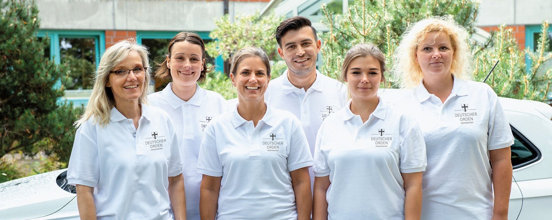 Gruppenfoto: Pfleger/innen in weissen DO-Shirts vor einem weissem Kleinwagen