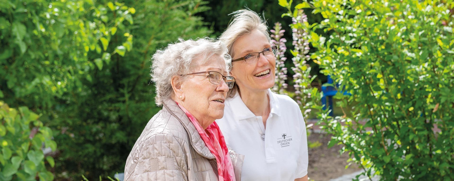 Eine Pflegerin zeigt einer Bewohnerin etwas im Garten (nicht sichtbar) - beide lächeln