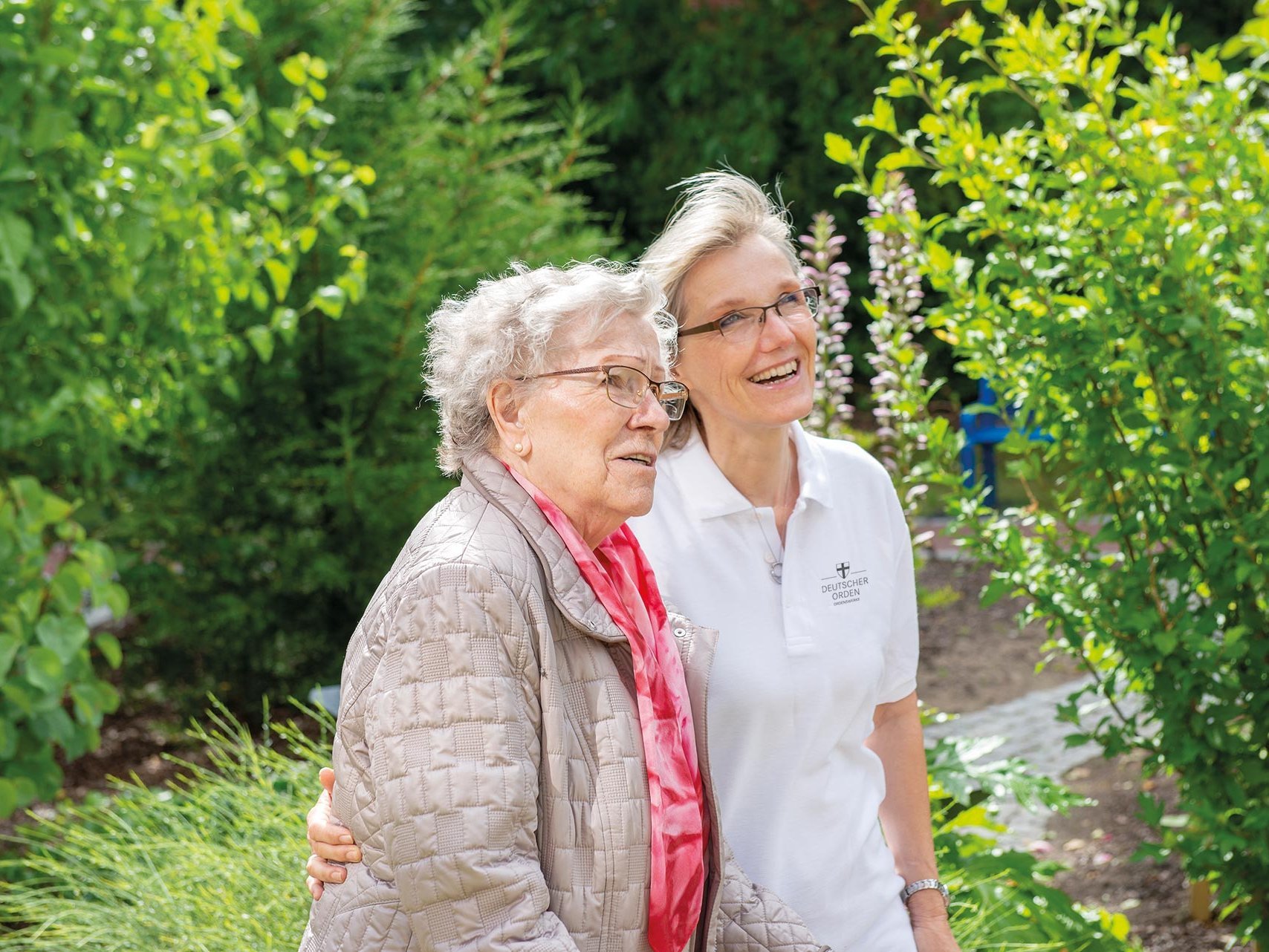 Eine Pflegerin zeigt einer Bewohnerin etwas im Garten (nicht sichtbar) - beide lächeln